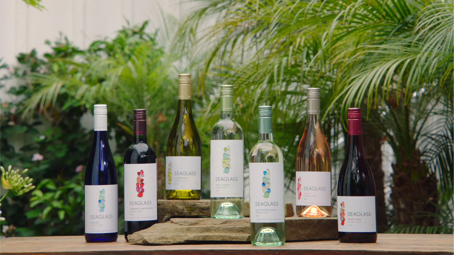 Seven bottles of Seaglass central coast wine, arranged on a table with palm trees in the background
