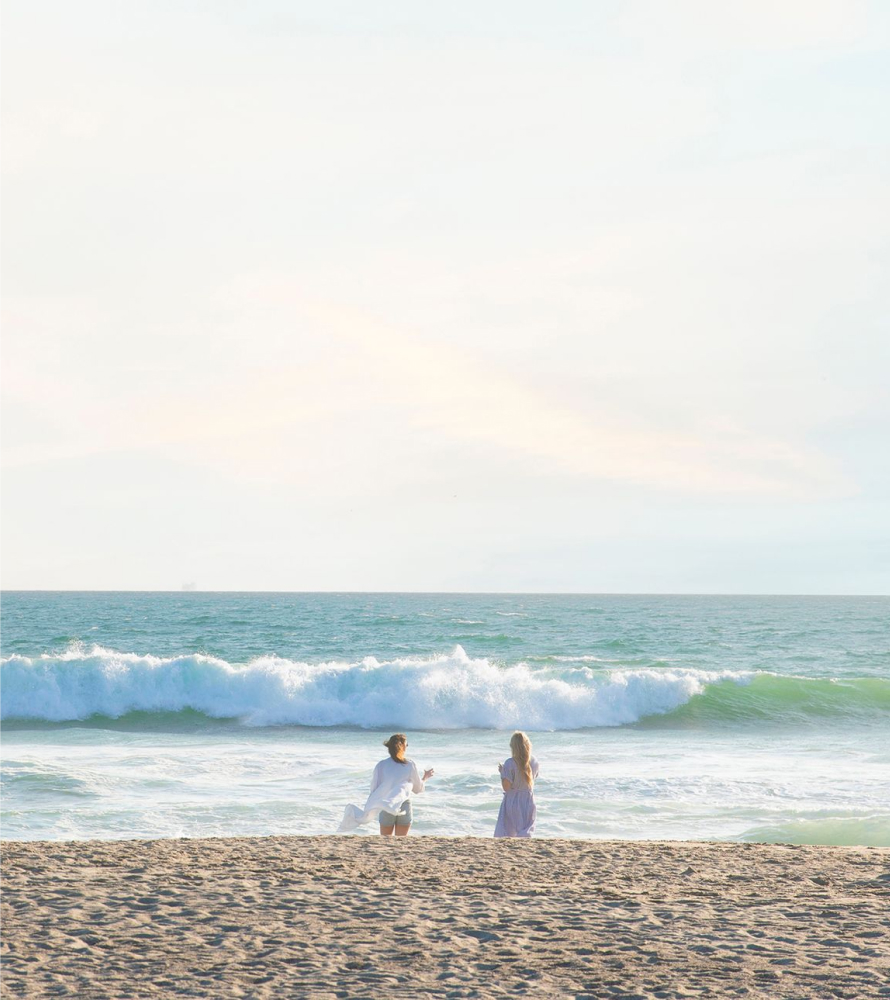 Two women stand on the water's edge in the middle distance with a sandy beach in the foreground and crashing ocean waves in the background