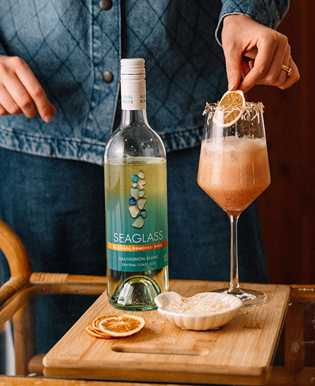 Someone adds a garnish to a tall glass of coconut blood orange mocktail next to a bottle of Seaglass alcohol-removed Sauvignon Blanc