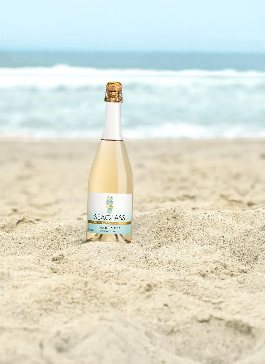 A bottle of SEAGLASS Coastal Wines Alcohol Removed Sparkling Brut in the sand on the beach